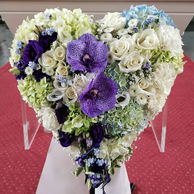 Hjerteformet blomsteroppsats i hvitt, blått og lilla med to store orkideer i midten.