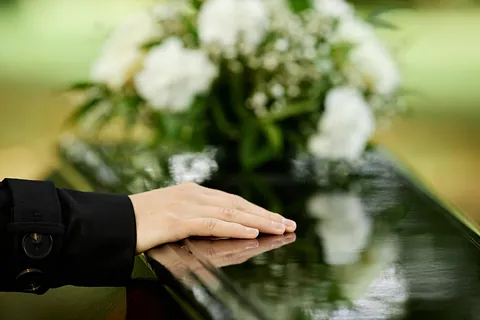 Nærbilde av hånd som hviler på blank kistelokk med hvite blomster i bakgrunnen.