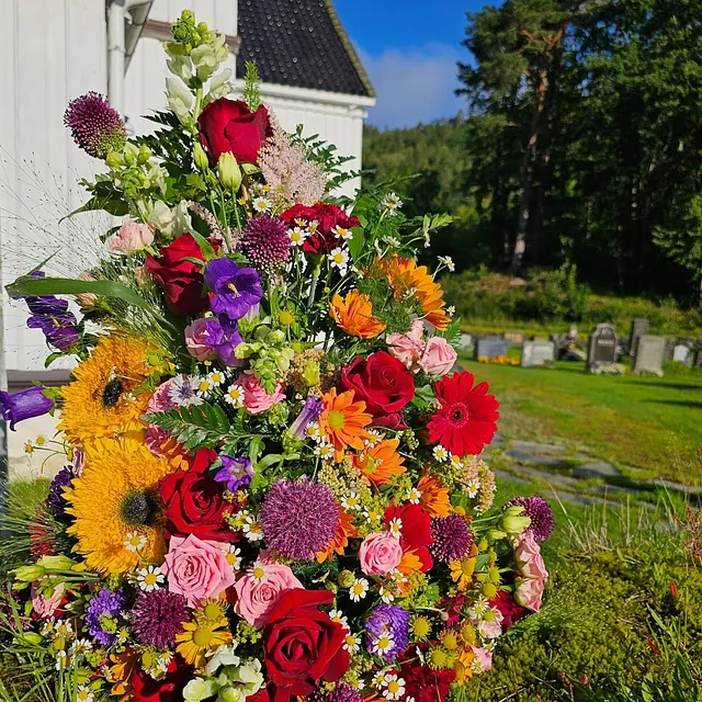 Fargerik blomsteroppsats med roser, solsikker og markblomster plassert ute ved kirke og gravplass.