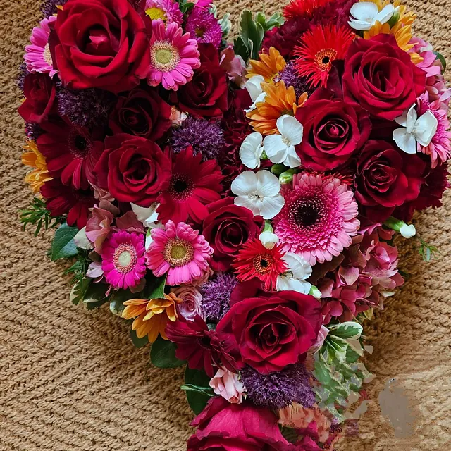 Hjerteformet blomsteroppsats med røde roser, rosa germini og fargerike innslag mot grov bakgrunn.
