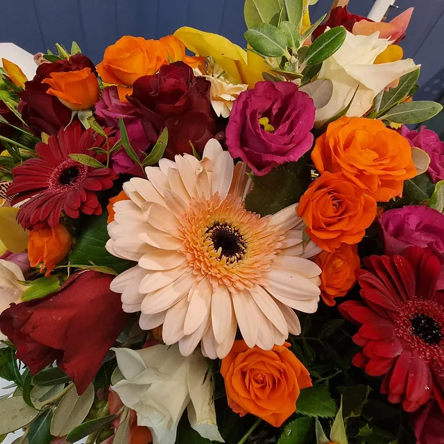 Fargerik blomsterbukett med lys gerbera i midten, omgitt av roser, nelliker og grønne blader.